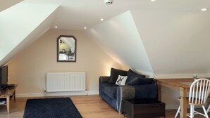 Un espace de vie de style loft avec un plafond en pente, un canapé noir avec des coussins, une table à manger en bois, une chaise blanche, un tapis noir et un radiateur blanc.