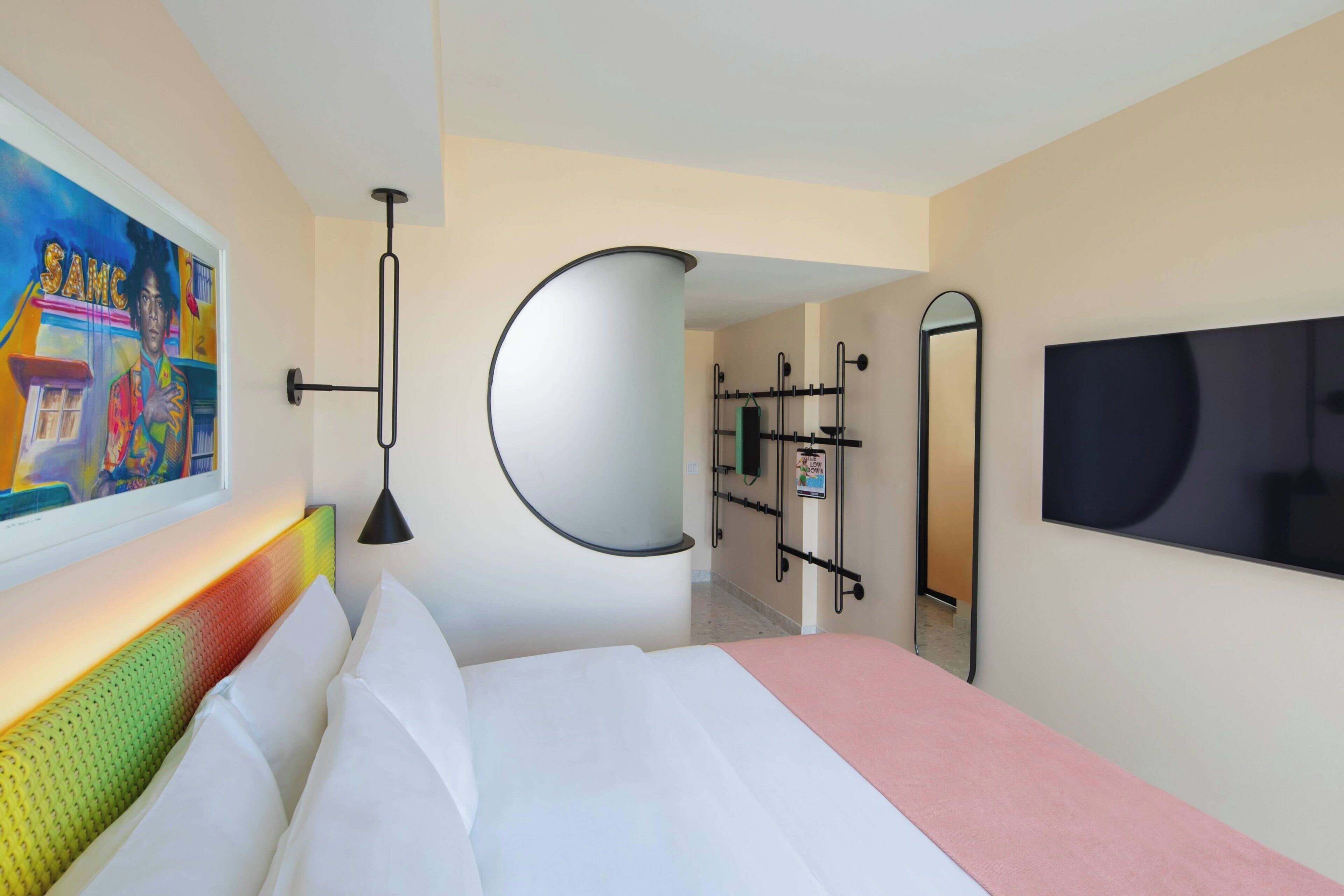 A hotel room with a bed, a TV, a painting, a mirror, and a bathroom with a shower and toilet.