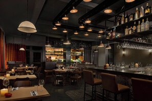A restaurant with multiple dining tables, hanging pendant lights, and a bar area with shelves of bottles.