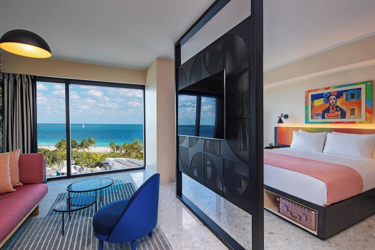 A hotel room with a large window offering a view of the ocean, a bed with a colorful headboard, a small table with chairs, and a sofa.
