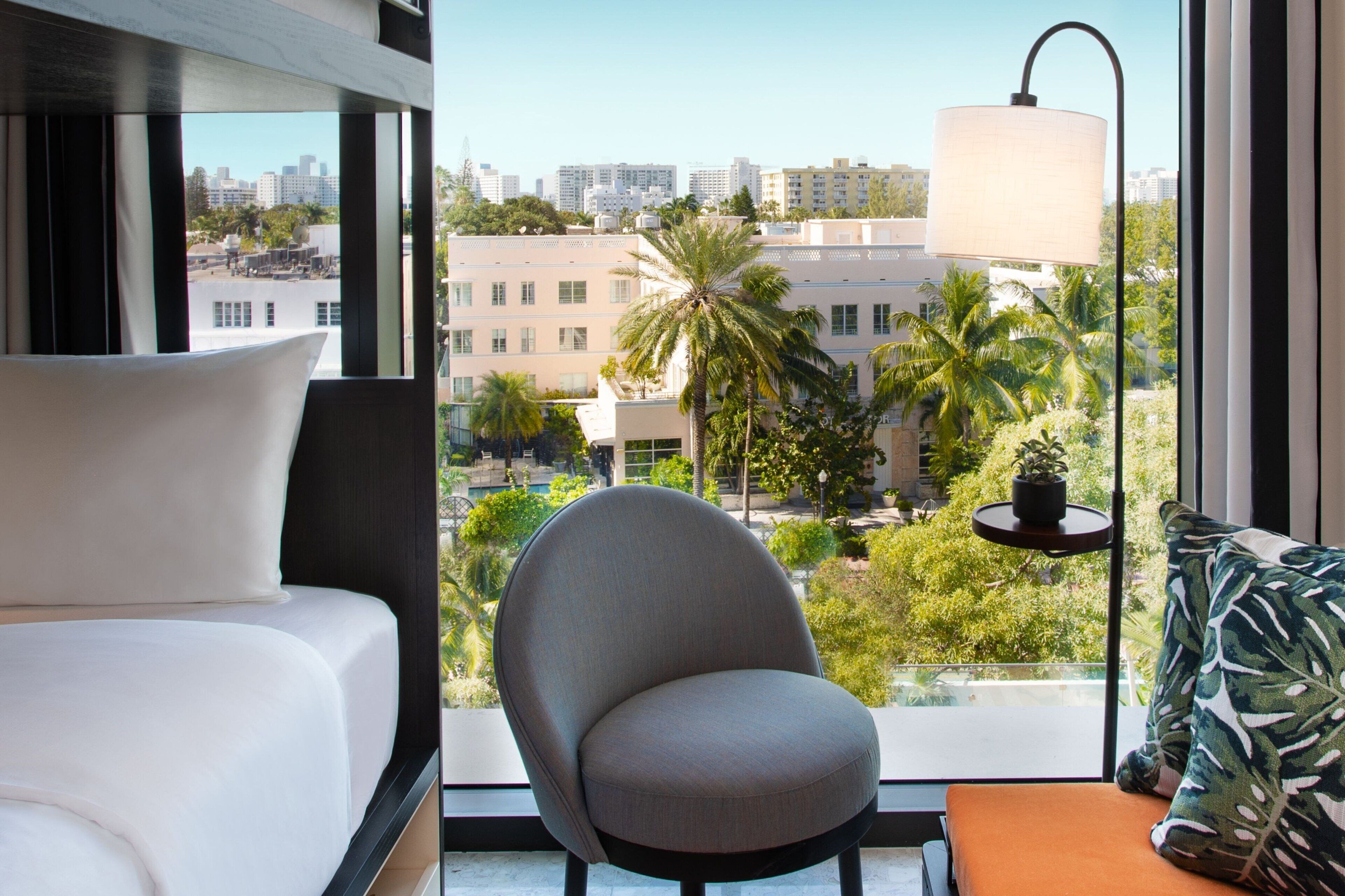 A modern hotel room with a large window offering a city view, a comfortable chair, a bedside table with a lamp, and a bed with white linens.
