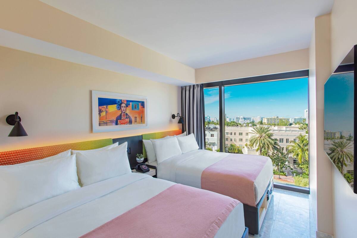 A hotel room with two beds, a large window with a city view, a painting on the wall, and a ceiling light.