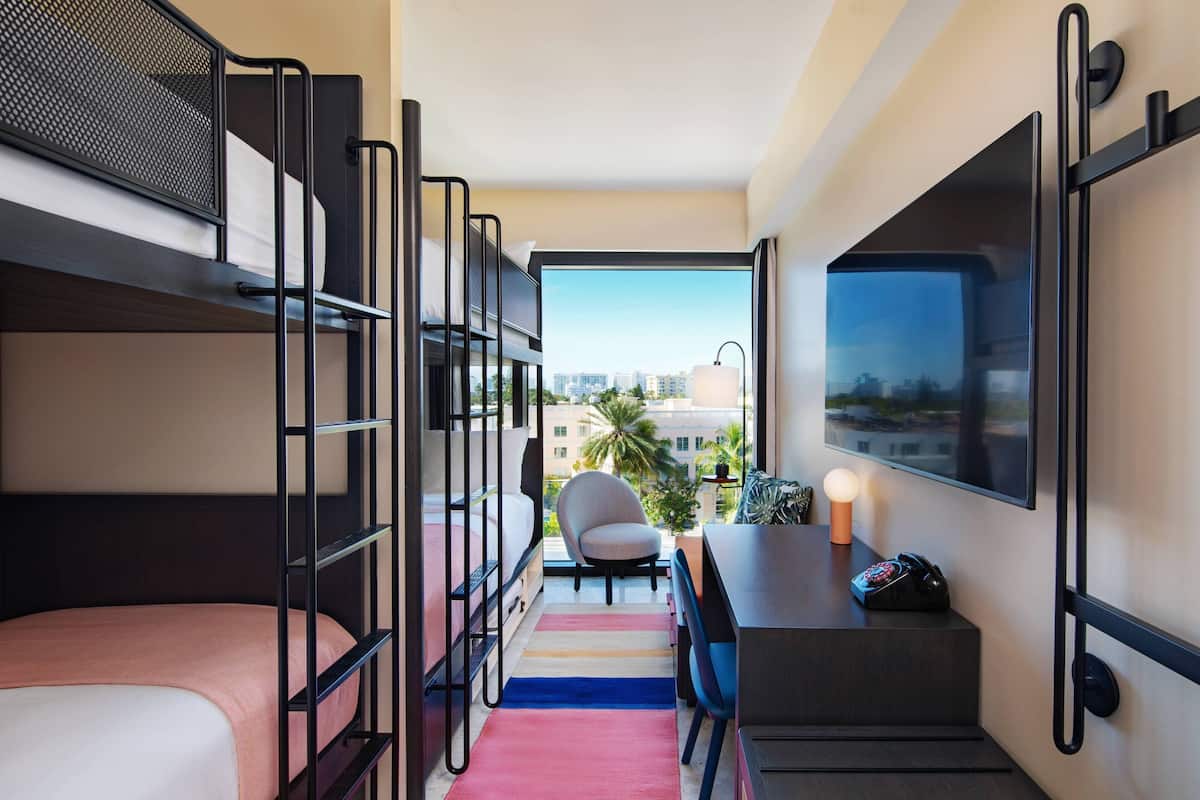 A compact hotel room with bunk beds, a desk, a chair, and a large window offering a city view.