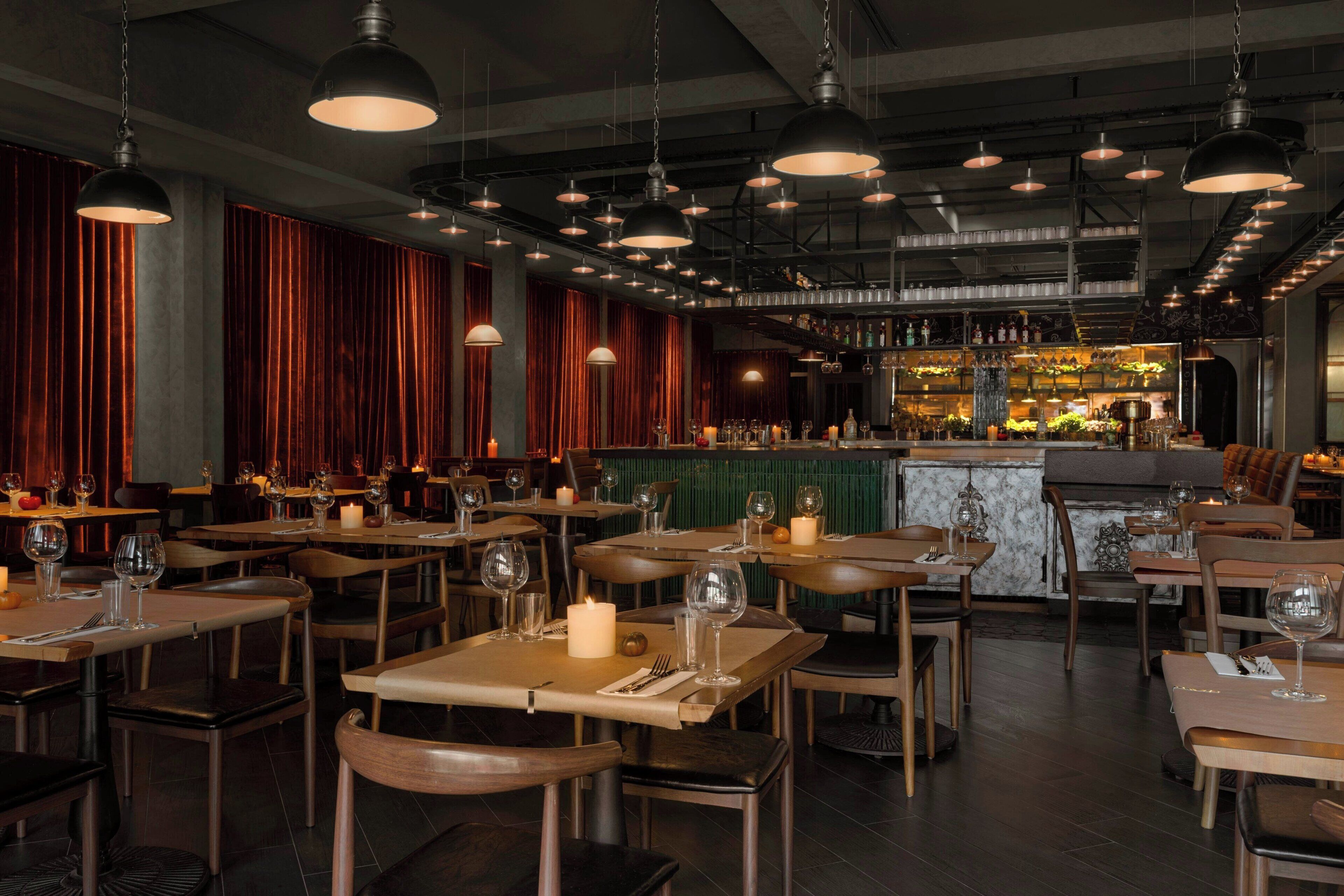 L’intérieur d’un restaurant présente plusieurs tables, des chaises, des luminaires suspendus, un bar avec des bouteilles exposées et un comptoir vert.