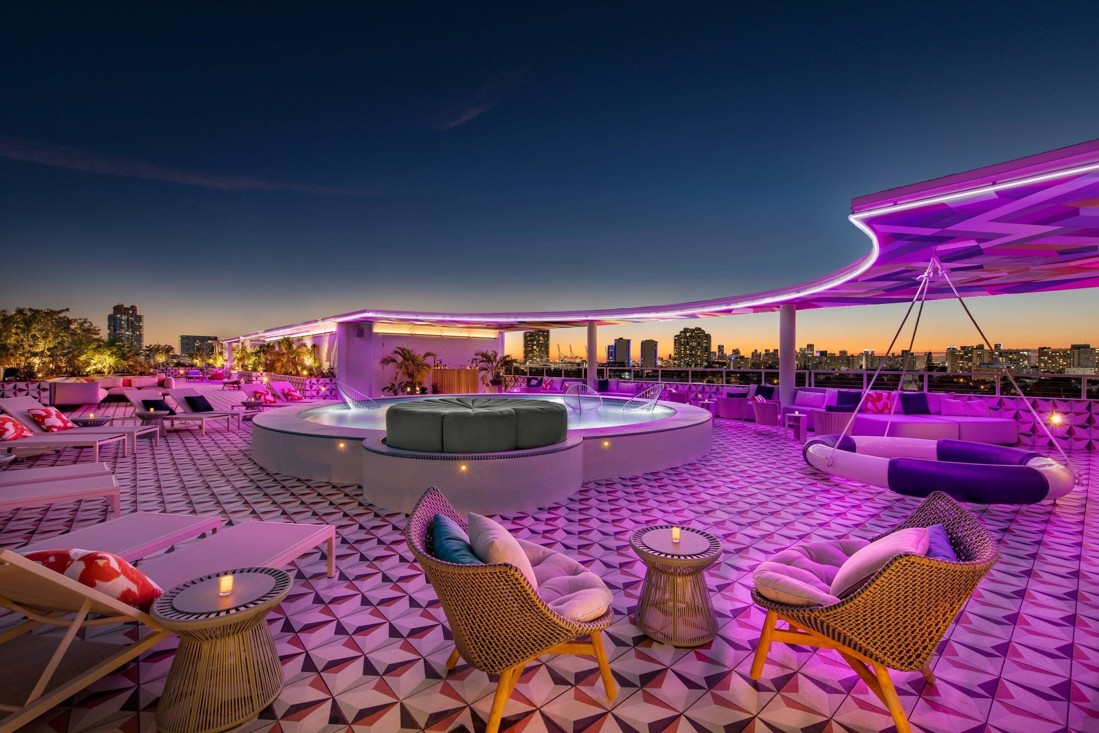 Un salón en la azotea con piscina, sillones de descanso y vistas a la ciudad al anochecer.