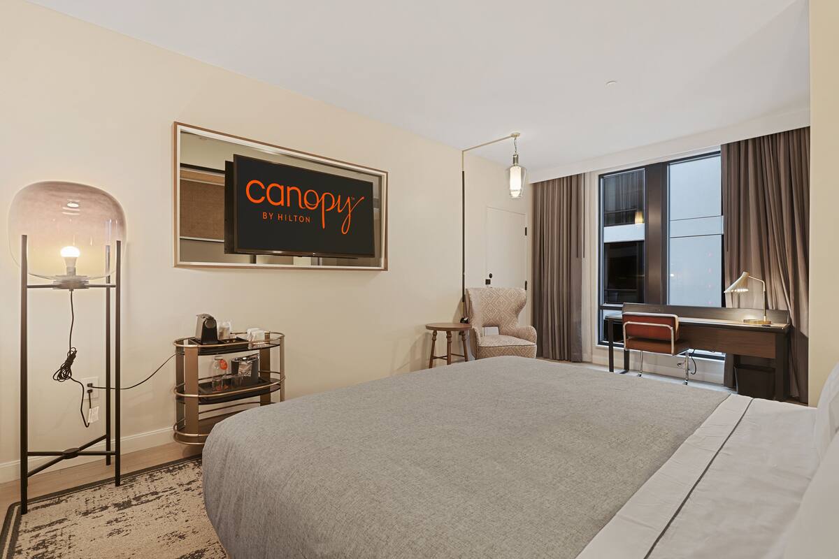 Une chambre d’hôtel avec un grand lit, un bureau avec une chaise, une lampe sur pied, un téléviseur fixé au mur et une fenêtre avec des rideaux.