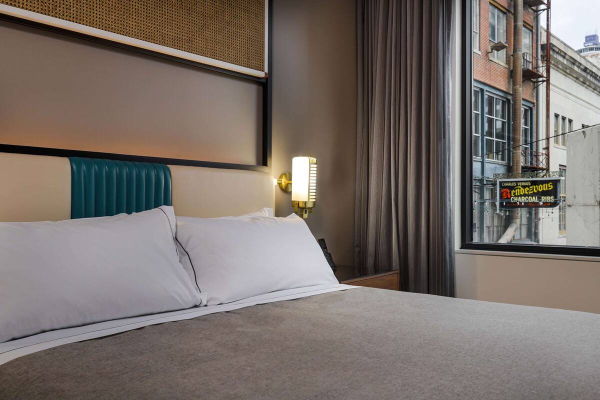 Une chambre d’hôtel avec un lit, une tête de lit, une lampe fixée au mur et une vue sur la rue.