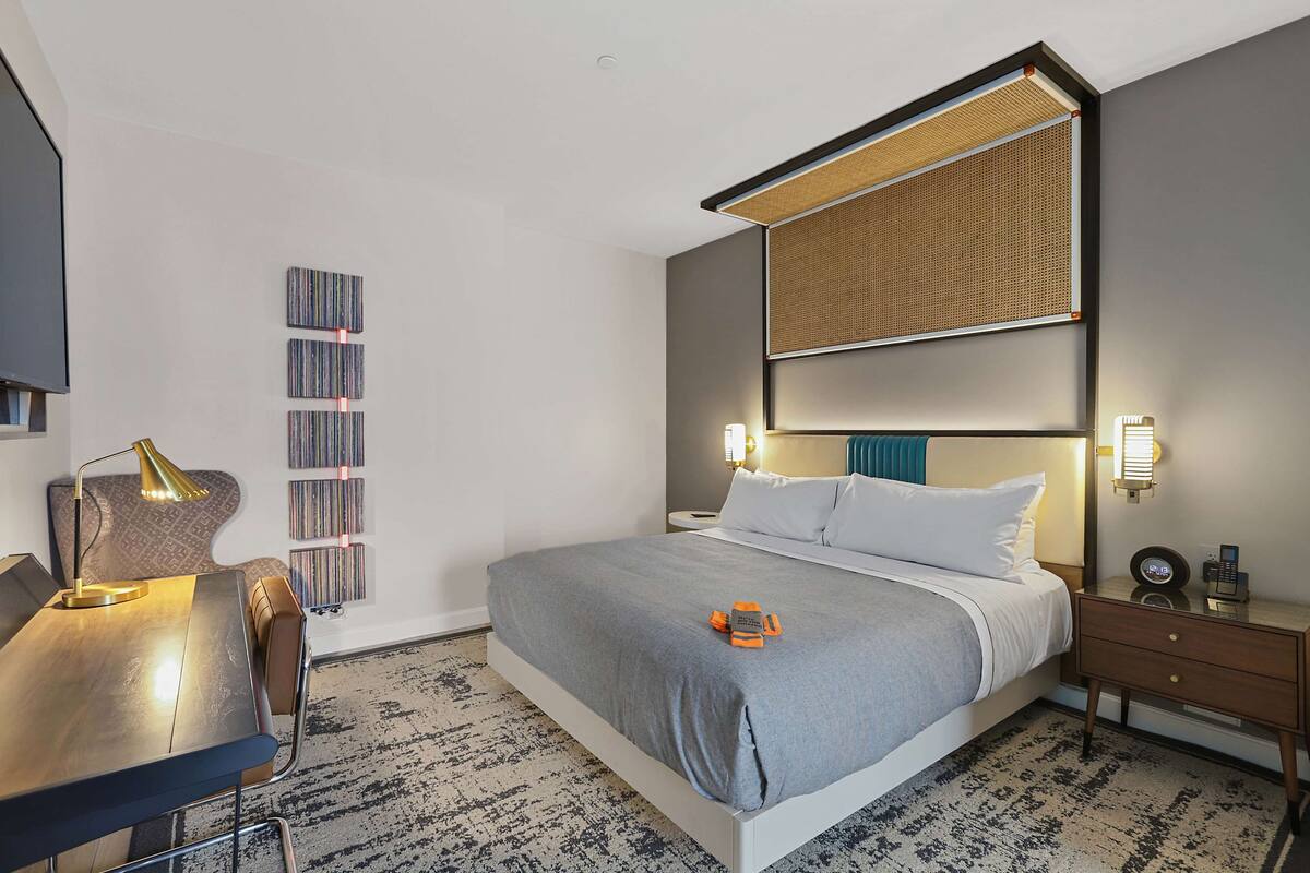 Une chambre d’hôtel moderne dotée d’un grand lit, d’un bureau avec une lampe, d’une bibliothèque et d’une grande fenêtre offrant une vue dégagée.