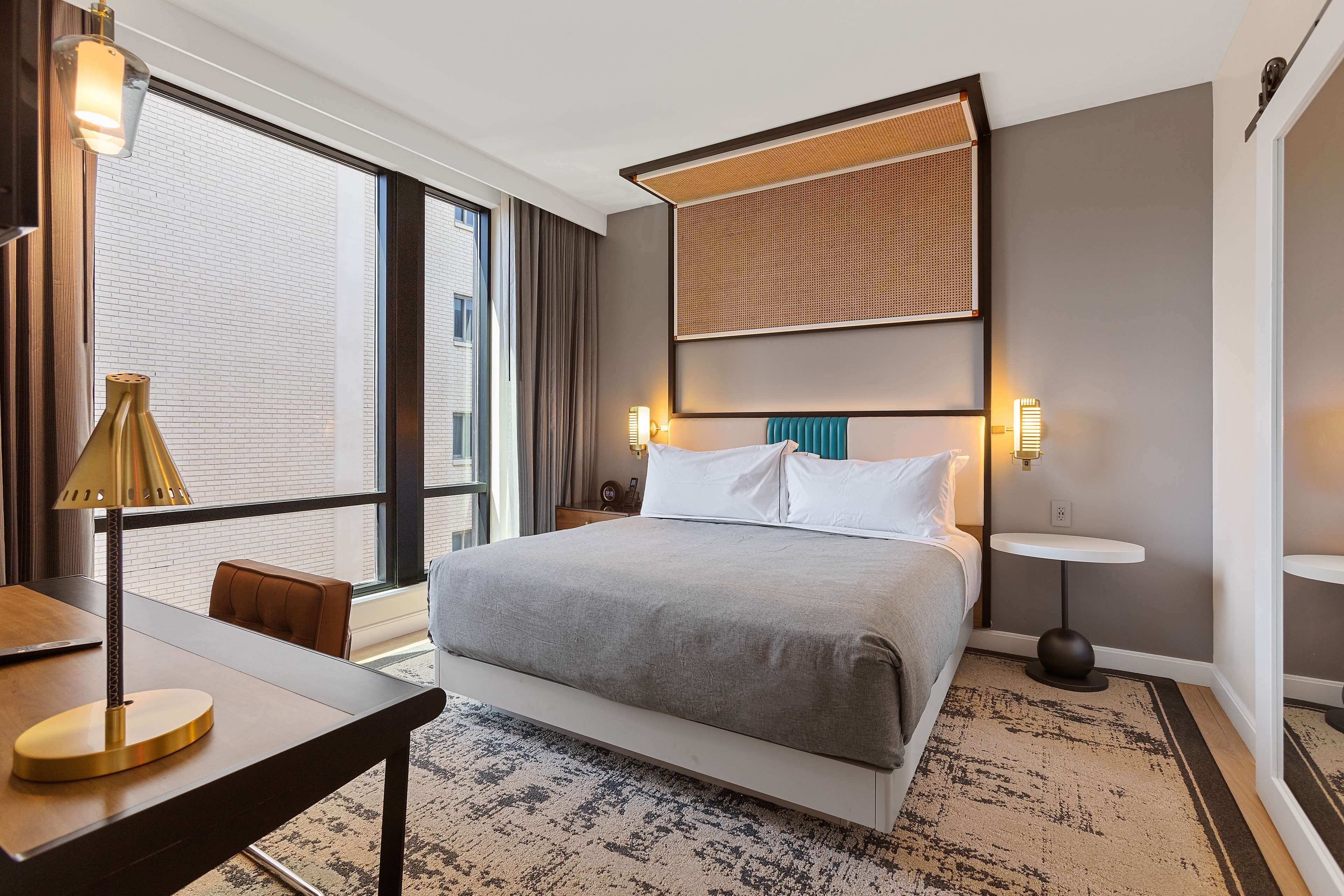 A modern hotel room with a large bed, a desk, a chair, a small table, and a large window with curtains.