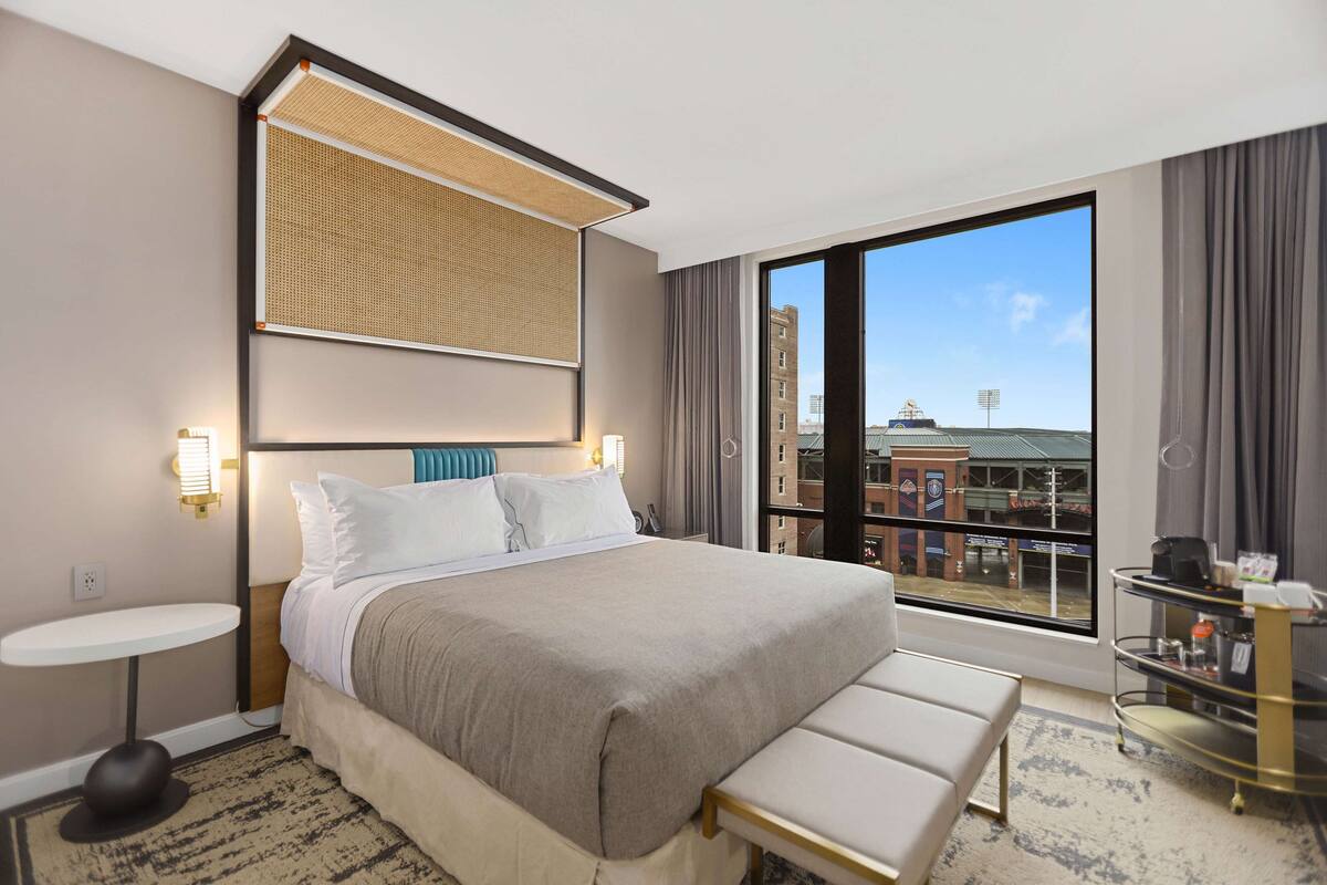 Une chambre d’hôtel avec un grand lit, une petite table ronde, un banc, une fenêtre donnant sur des immeubles et un minibar.