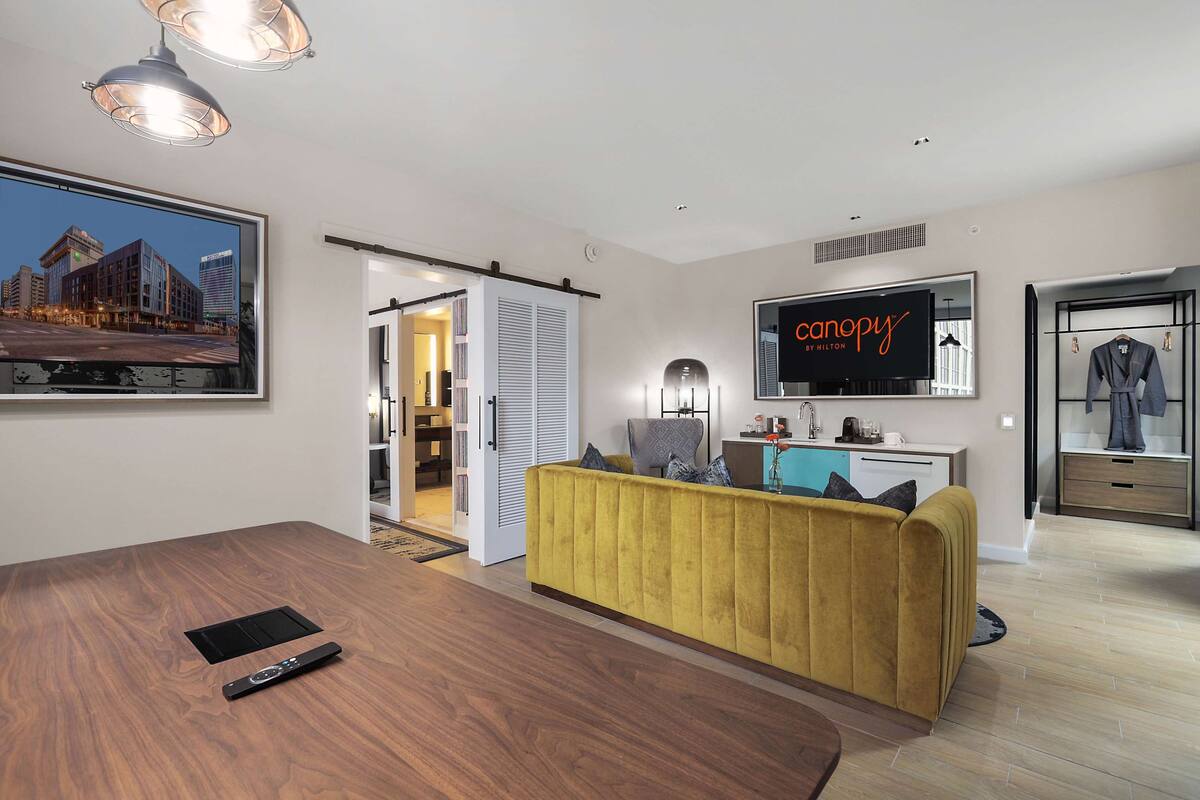 Un salon moderne avec un canapé jaune, une table à manger et un téléviseur diffusant l'émission « Caropy by Hilton ».