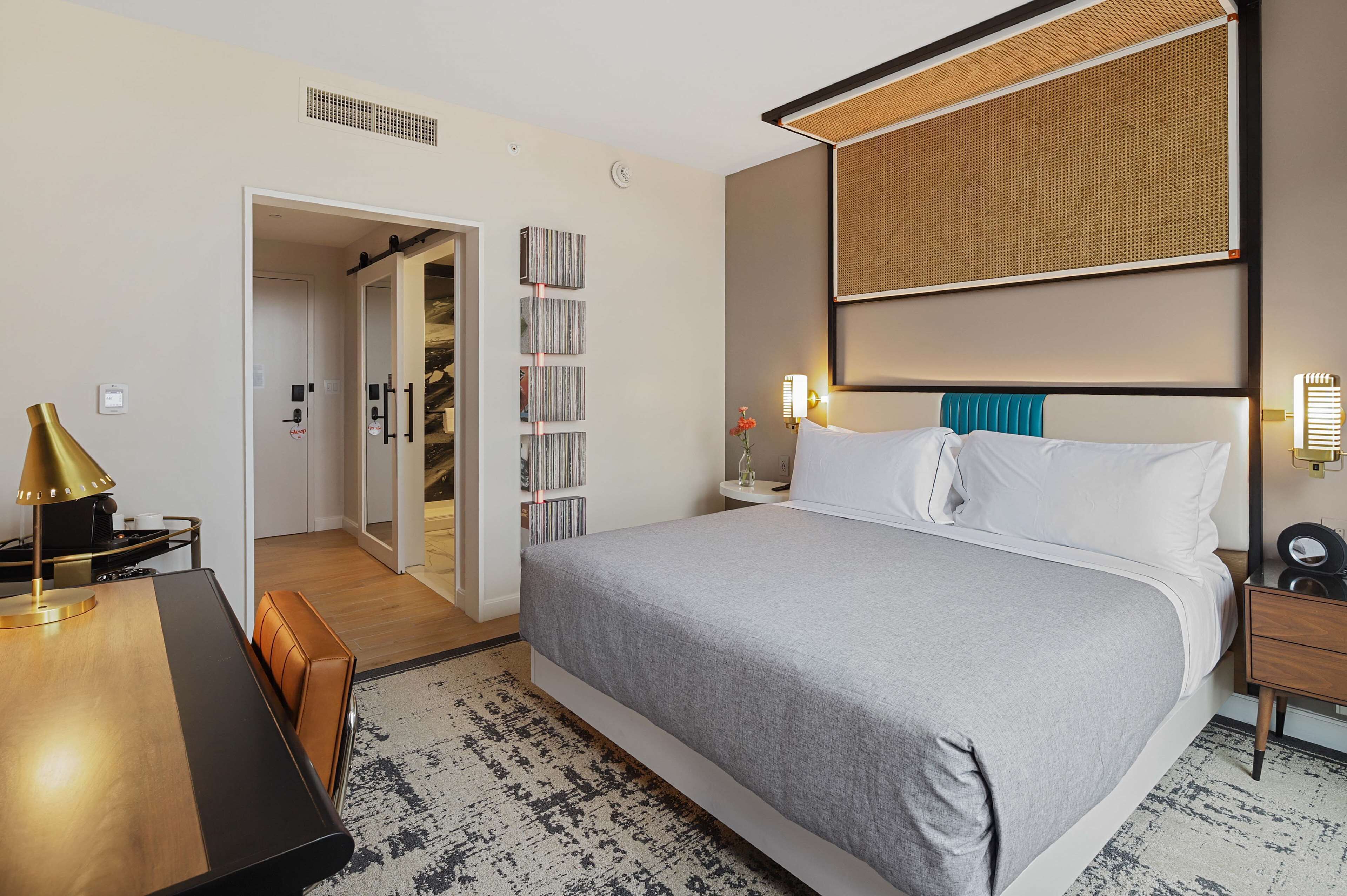Une chambre d’hôtel avec un grand lit, un bureau avec une lampe, une bibliothèque et une salle de bain visible à travers une porte ouverte.