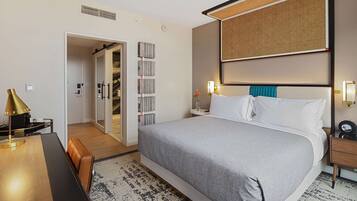 Une chambre d’hôtel avec un grand lit, un bureau avec une lampe, une bibliothèque et une salle de bain visible à travers une porte ouverte.