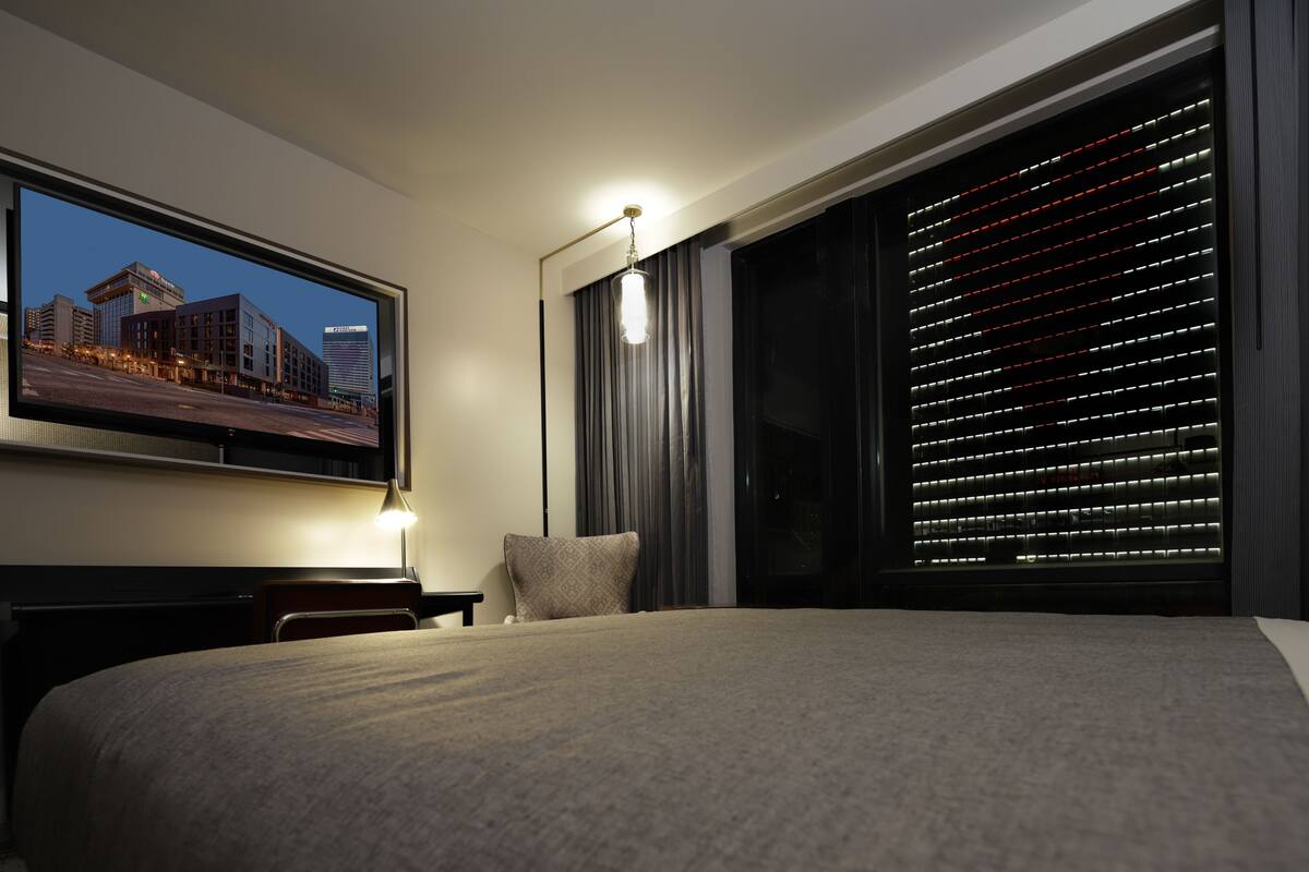 Une chambre d’hôtel avec un grand lit, un téléviseur à écran plat fixé au mur, une table de chevet, une lampe et une fenêtre avec des stores.