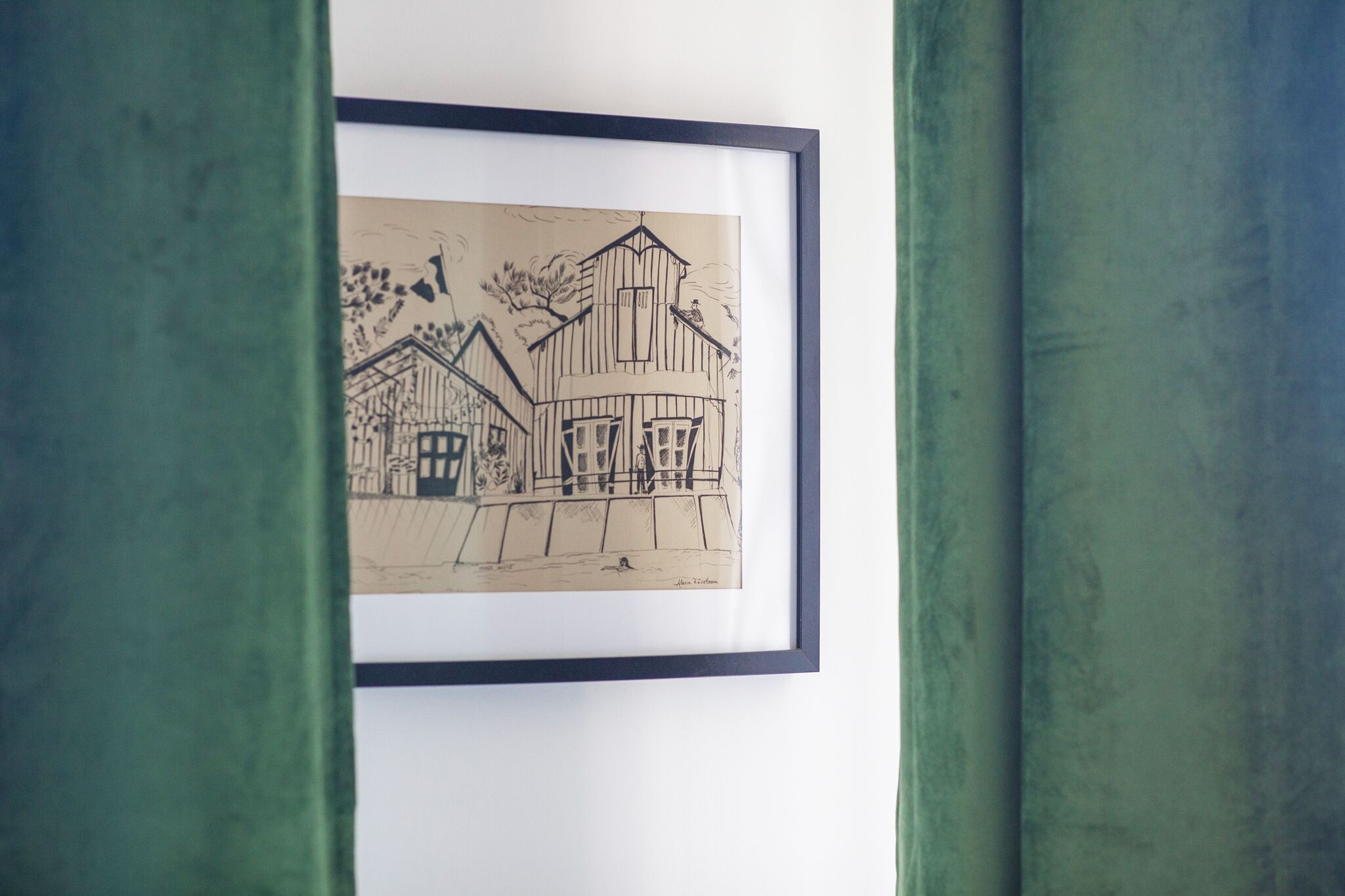 A framed black and white drawing of a building with a gabled roof and a chimney, mounted on a white wall.