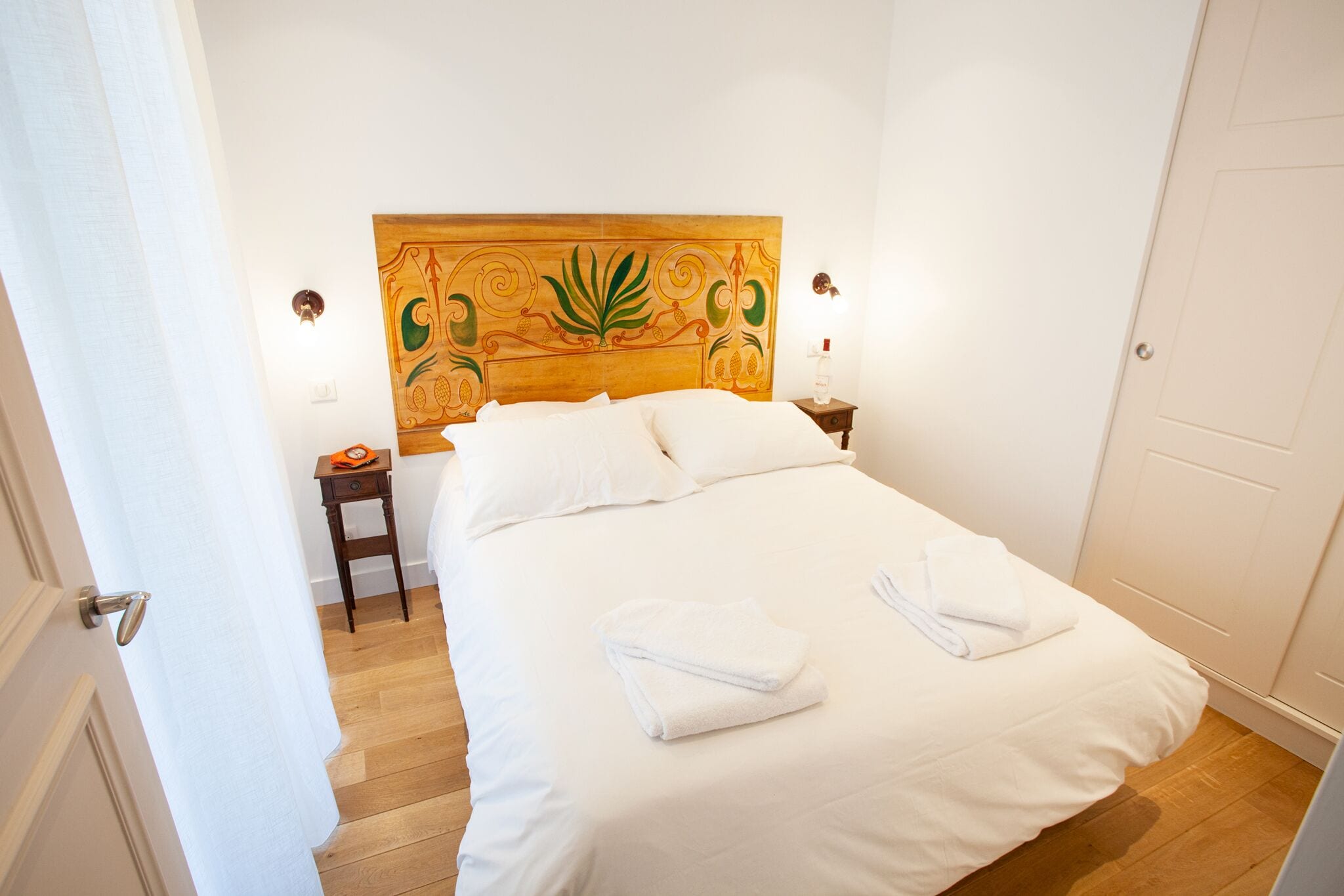 Un lit bien fait, avec du linge de lit blanc et deux serviettes pliées, une tête de lit en bois ornée d’un motif de feuilles et une petite table de chevet avec une lampe.