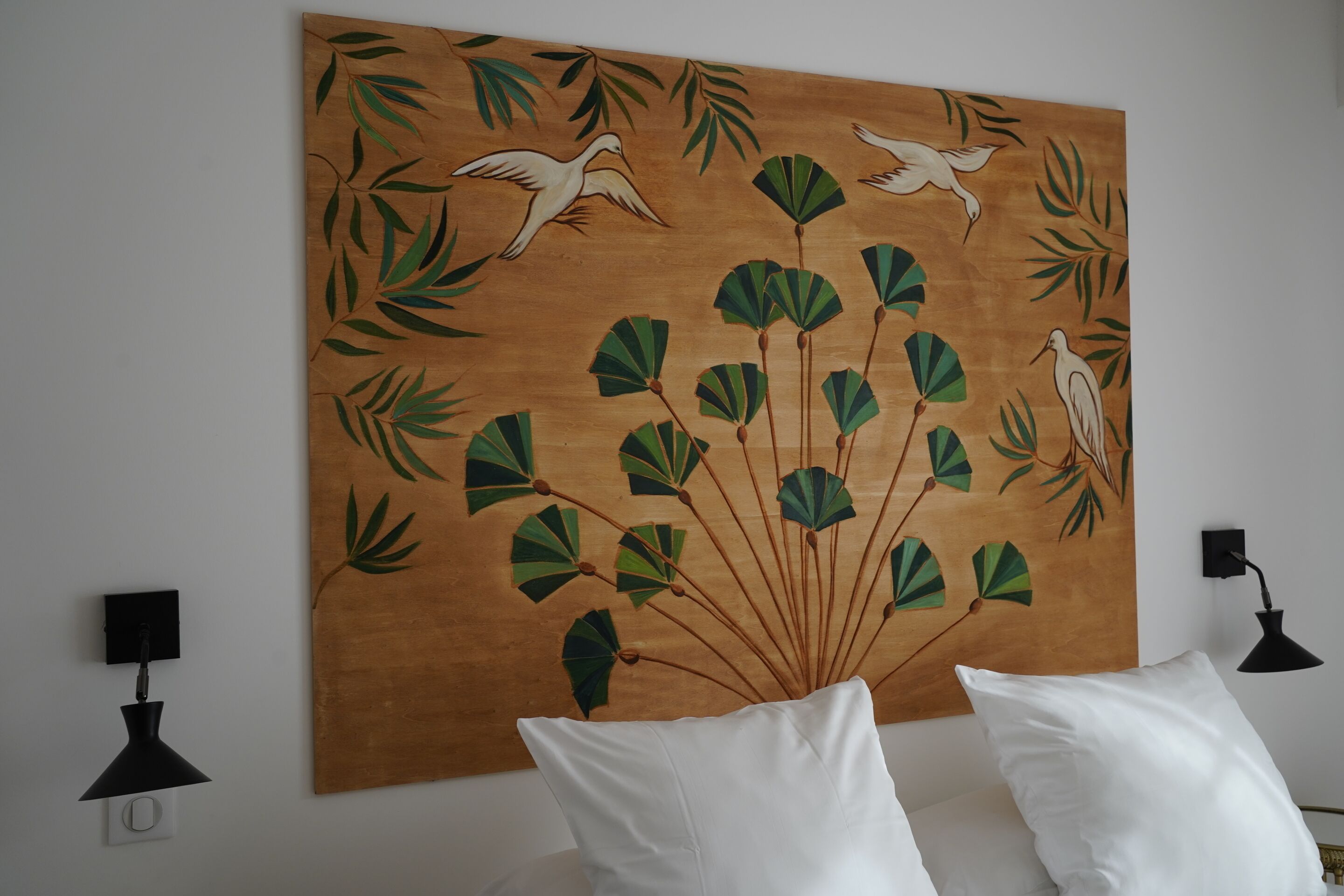 Een kamer met een groot muurschilderij van vogels en bladeren, witte kussens en zwarte wandlampen.