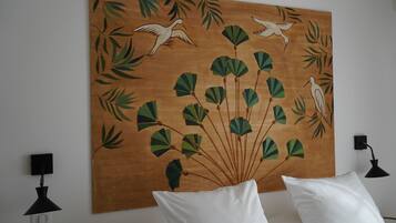 Een kamer met een groot muurschilderij van vogels en bladeren, witte kussens en zwarte wandlampen.
