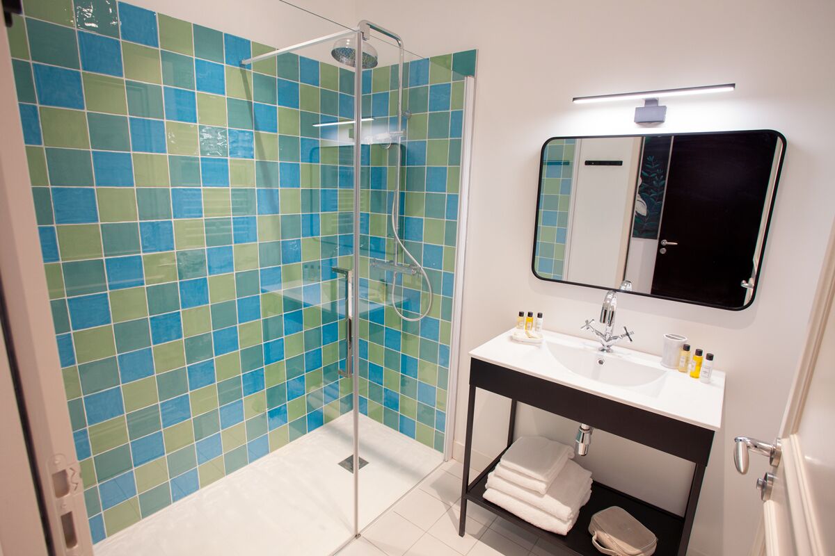 Ein Badezimmer mit einer gläsernen Duschkabine, einem weißen Waschbecken mit Spiegel darüber und einer gefliesten Wand mit blau-grünen Fliesen.