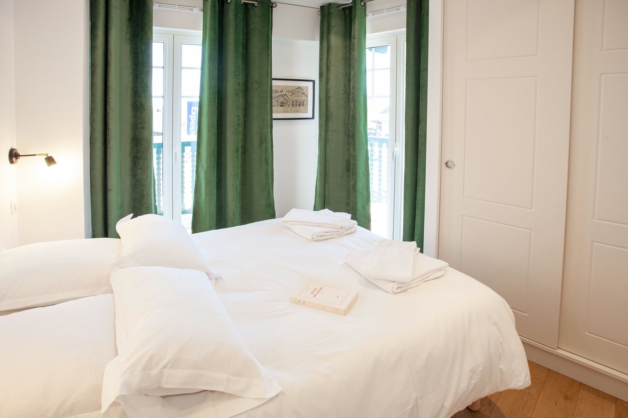 Habitación de hotel con dos camas, cortinas verdes, un cuadro en la pared y un libro sobre la cama.