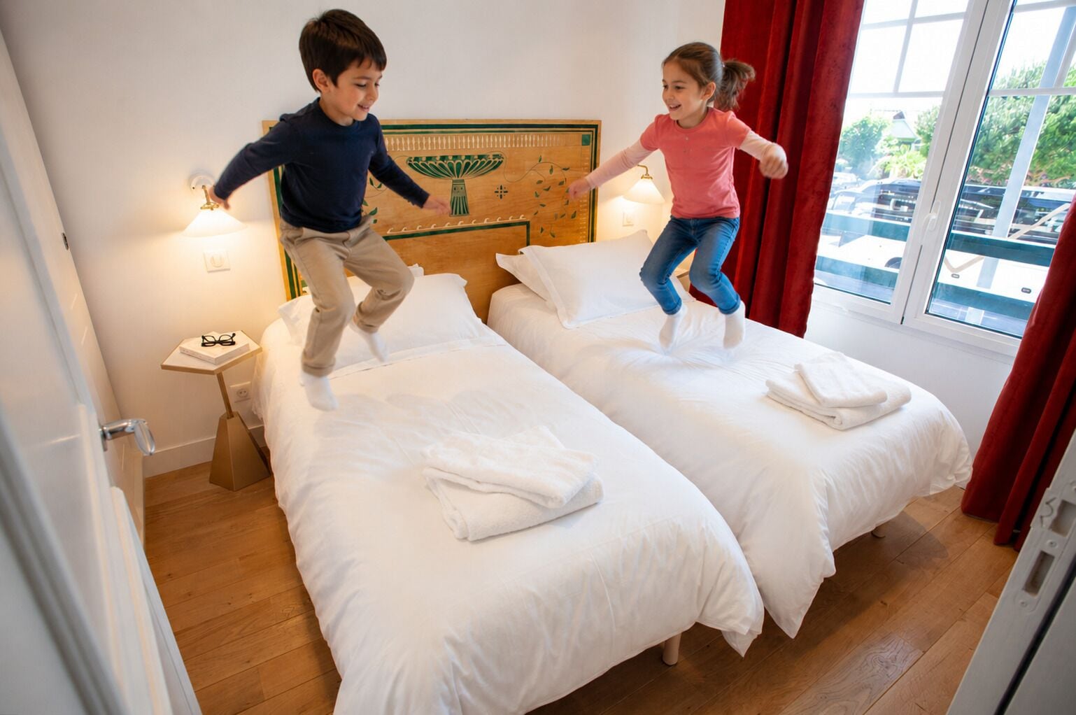 A hotel room with two beds, a wooden headboard, a nightstand, and a window with a view of the outside.