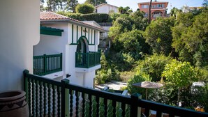 A balcony with a view of a lush garden, a white building with green accents, and a red building in the background.