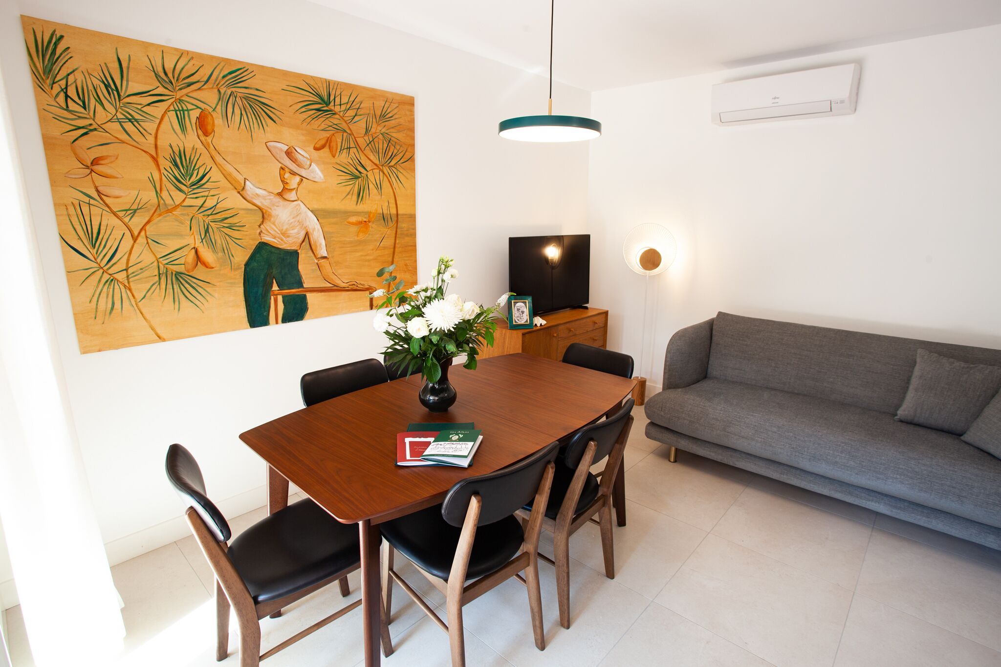 A dining area with a wooden table, black chairs, a grey sofa, a painting, a TV, and a vase with flowers.
