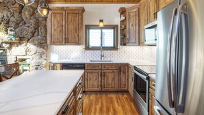 A kitchen with wooden cabinets, a stone wall, a stainless steel refrigerator, and a window with blinds. - Dog Friendly, On Shuttle Route, Private Hot Tub, Near River, Walk to Town (Breckenridge)