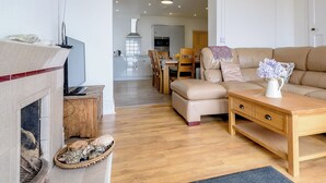 A living room with a fireplace, a sofa, a dining table with chairs, and a kitchen in the background. - Beachside, West Yardley House - UK31200 (Downderry and Seaton, near Looe)