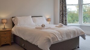 A neatly made bed with white linens and two rolled towels, a bedside table with a lamp, and a window with curtains. - 4 bedroom accommodation in Keswick (Keswick)