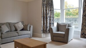 A living room with a grey sofa, a matching armchair, and a wooden coffee table. There is a large window with a view of trees and a stone wall outside. - 4 bedroom accommodation in Keswick (Keswick)