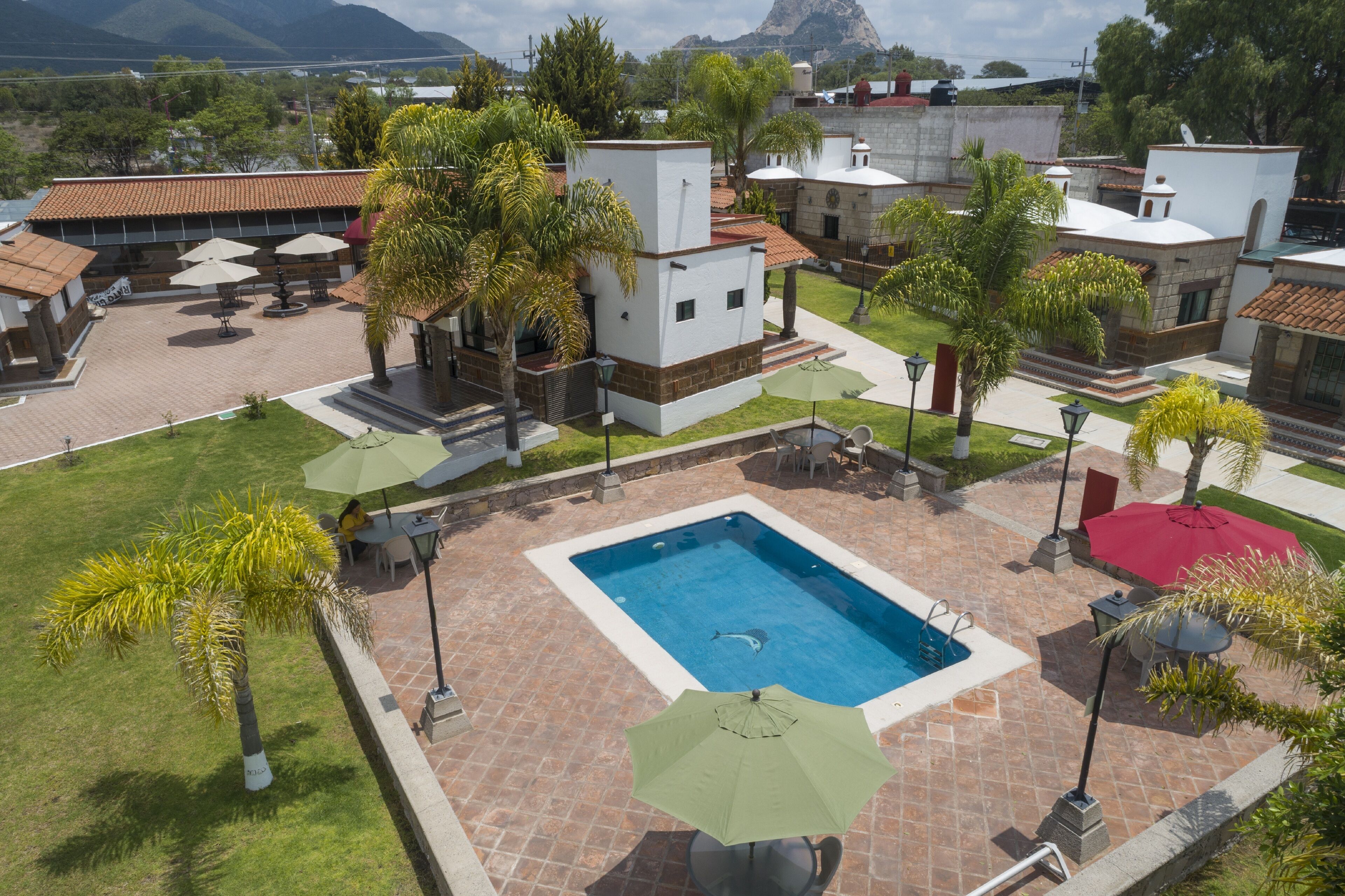 Zona de piscina con una pileta rectangular, rodeada de palmeras y sombrillas, y contigua a un edificio con techo de tejas.
