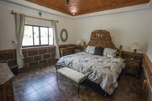 Un dormitorio con una cama grande, un banco, una ventana con cortinas, un cabecero de madera y una pequeña mesa con una lámpara.