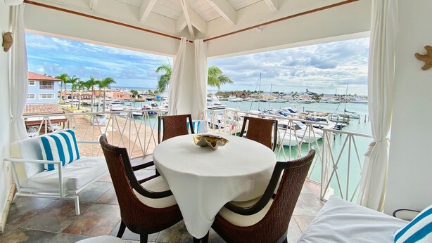 Un espacio de estar techado al aire libre con una mesa y sillas, con vista a un puerto deportivo con barcos y edificios.