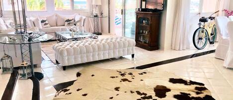 Un salón amplio con una gran mesa de centro de vidrio, un sofá blanco capitonado, un tapiz de cuero de vaca y un televisor sobre un mueble de madera.