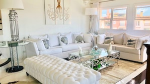 Un salón moderno con un sofá blanco, una mesa de centro de vidrio y un otoman tapizado.
