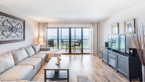 Un salon moderne avec un canapé, une table basse et un téléviseur posé sur un meuble. Il y a un balcon avec vue sur la mer.