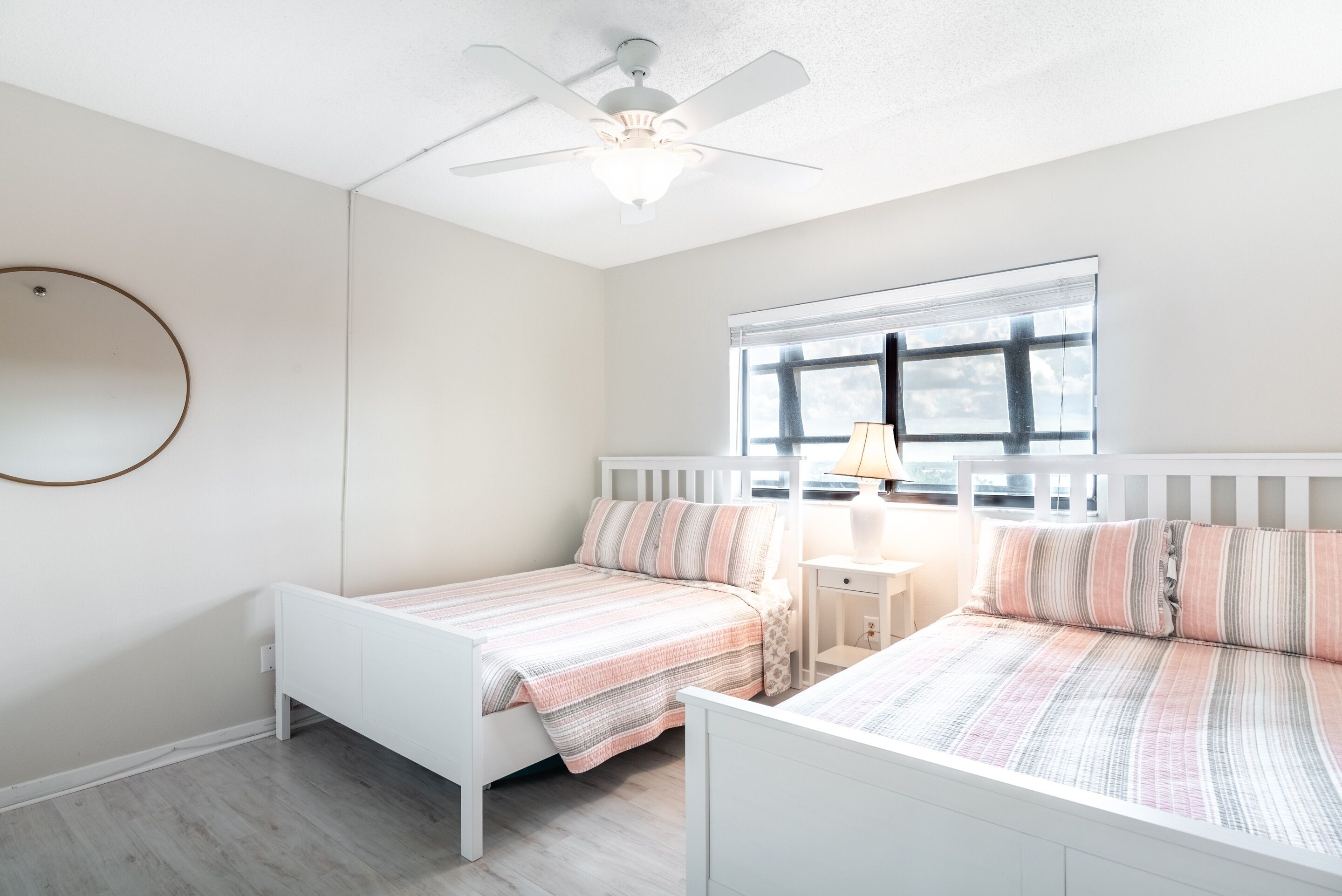 A bedroom with two beds, a ceiling fan, a window with blinds, and a bedside table with a lamp.