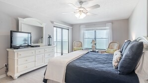 Une chambre à coucher comprenant un lit, une commode, un téléviseur, un ventilateur de plafond et de grandes fenêtres.