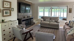 A living room with a fireplace, a television mounted on the wall, and a large window overlooking a lake. - Ultimate Family Lake Retreat w/ Swim Slide, Sandy bottom, and Bar/TV Lounge (Holcombe)
