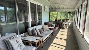 A covered porch with a row of wooden benches and cushions, a table with chairs, and large windows offering a view of the outdoors. - Ultimate Family Lake Retreat w/ Swim Slide, Sandy bottom, and Bar/TV Lounge (Holcombe)
