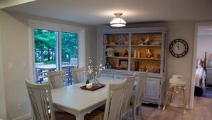 A dining area with a table and chairs, a window with a view of trees, a shelf with decorative items, and a clock on the wall. - Ultimate Family Lake Retreat w/ Swim Slide, Sandy bottom, and Bar/TV Lounge (Holcombe)