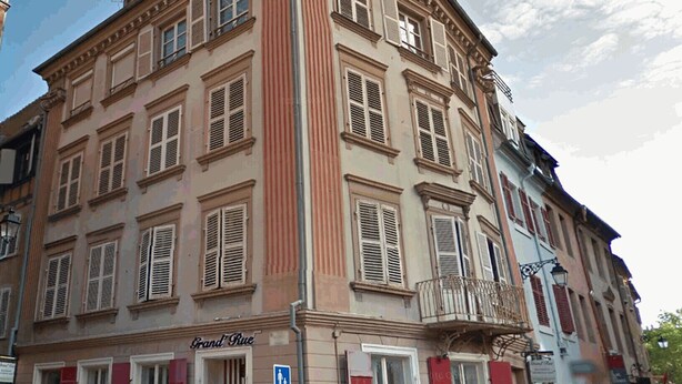 Ein historisches Gebäude mit mehreren Fenstern, einem Eingang im Erdgeschoss und einem sichtbaren Schild mit der Aufschrift 'Grand Rue'.