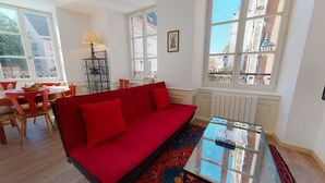 Ein Zimmer mit einem roten Sofa, einem Glastisch, einem Esstisch mit Stühlen und großen Fenstern mit Blick auf Gebäude.