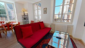 Une pièce avec un canapé rouge, une table basse en verre, une table à manger avec des chaises et de grandes fenêtres donnant sur des immeubles.