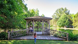 A wooden gazebo with a bench, surrounded by lush greenery and a pond. - The Holly House at Green Frog Farm (Alamo)