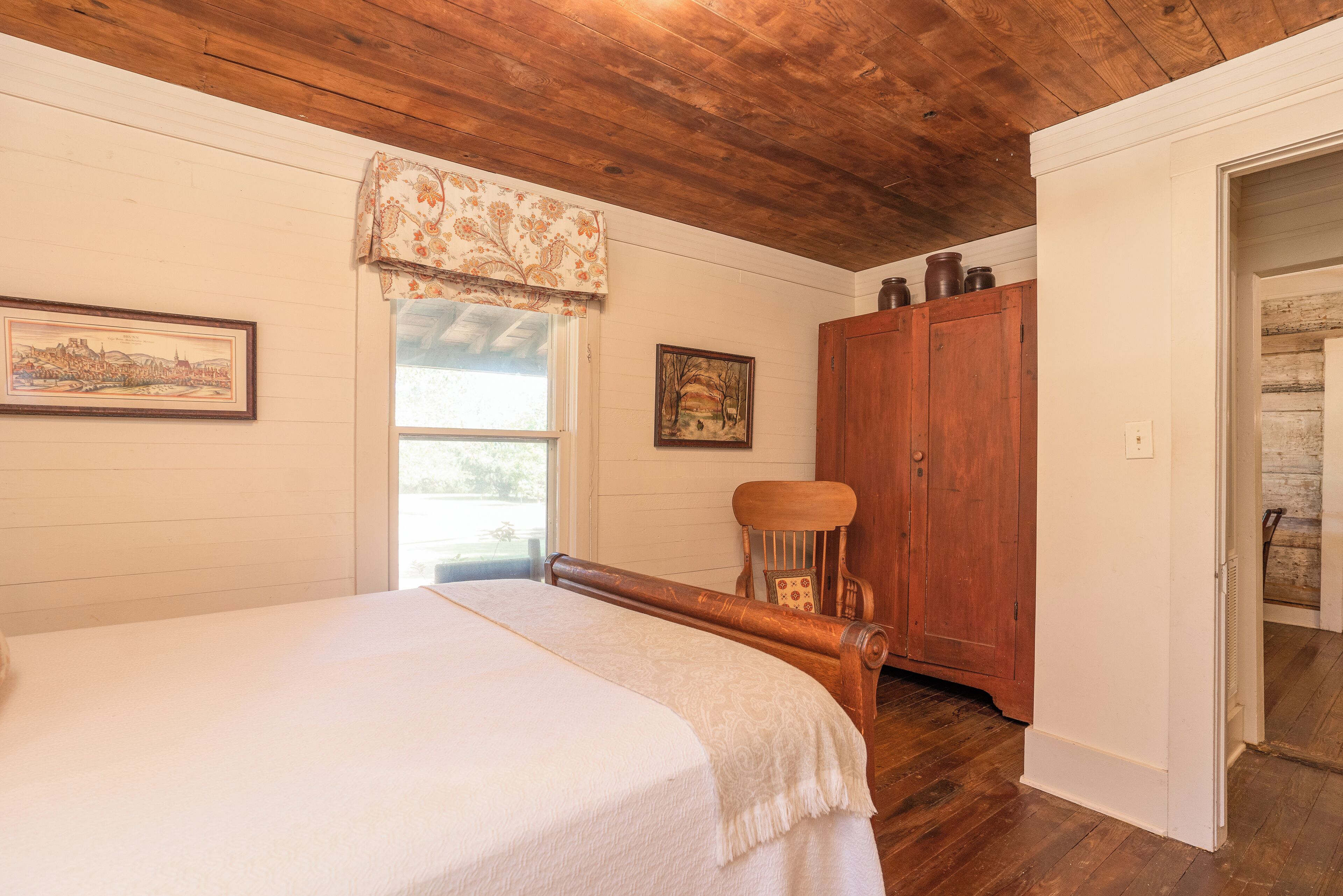 A bedroom with a wooden bed, a chair, a wardrobe, and a window with a floral valance.