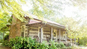 A rustic log cabin with a porch, surrounded by lush greenery and trees. - The Holly House at Green Frog Farm (Alamo)
