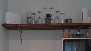 Et kompakt køkken med hvide skabe, en mikrobølgeovn, et lille køleskab og en træbordplade. Hylder overfor holder glas og krukker.