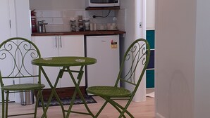 Et lille køkken med en mikrobølgeovn, hylder med glas og køkkenudstyr, samt et spiseområde med grønne stole og et bord.